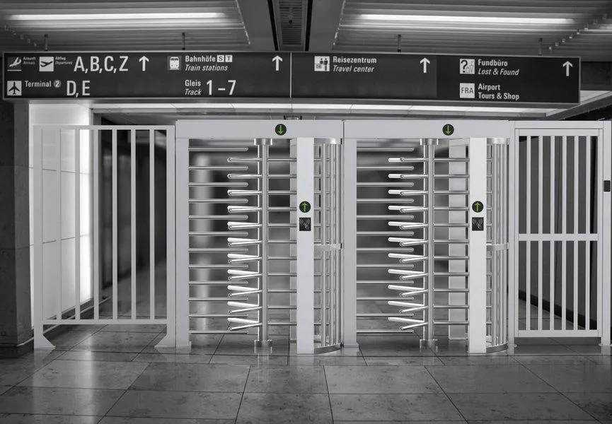 Dva celotelové prístupové turnikety DS411 z nehrdzavejúcej ocele, s bielymi vertikálnymi barmi a rotačnými ramenami, umiestnené vo vstupnej hale letiska alebo železničnej stanice, s informačnými tabuľami a navádzacími šípkami, zabezpečujúce kontrolu toku osôb.