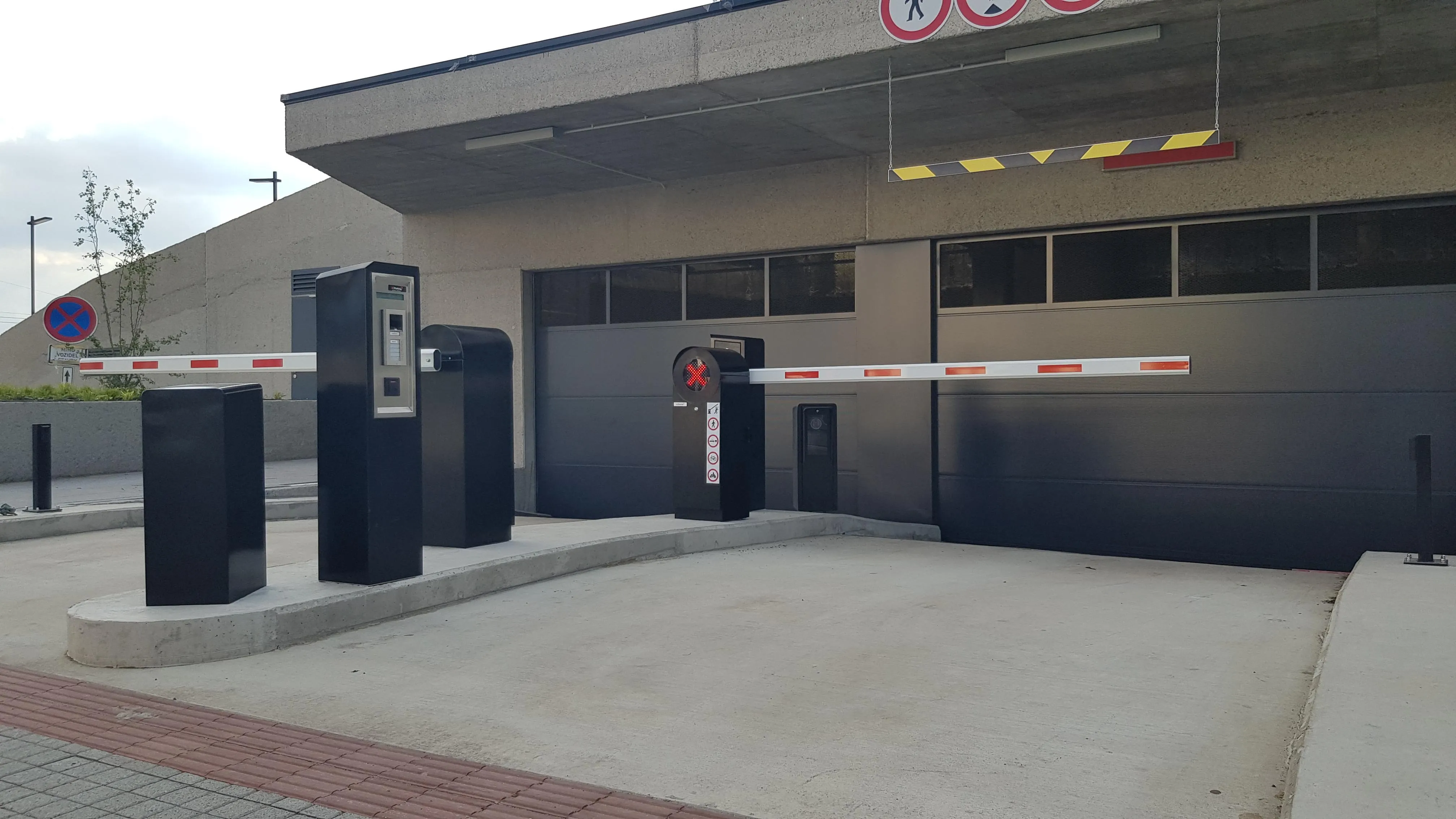 Vstup do podzemnej garáže s dvomi automatickými parkovacími závorami Park S v čiernej farbe s bielymi ramenami s červenými pruhmi, doplnenými o terminály pre platbu a riadenie prístupu, s tmavými garážovými dverami v pozadí.