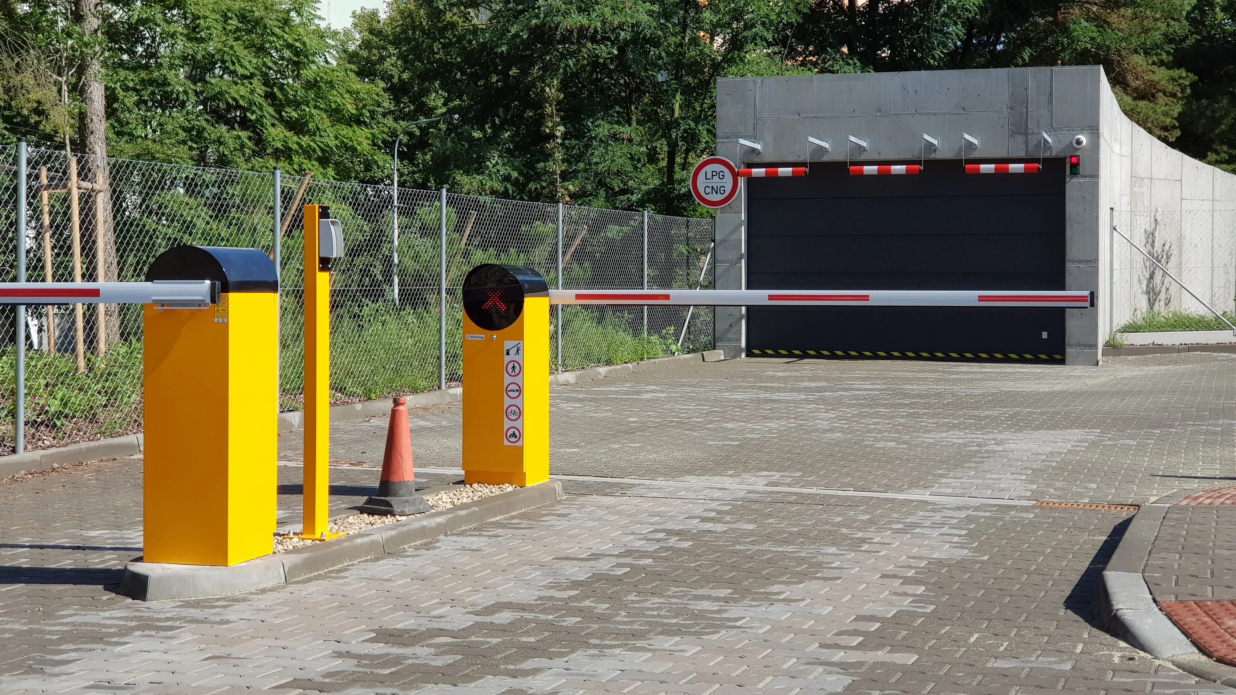 Vstup do podzemnej garáže s dvoma žltými automatickými závorami Park S, s bielymi ramenami s červenými pruhmi a signalizačnými svetlami, pričom ľavá závora má čierny vrchný kryt a pravá má integrovanú červenú stop značku, s tabuľkou "LPG CNG" a pletivovým oplotením po ľavej strane.
