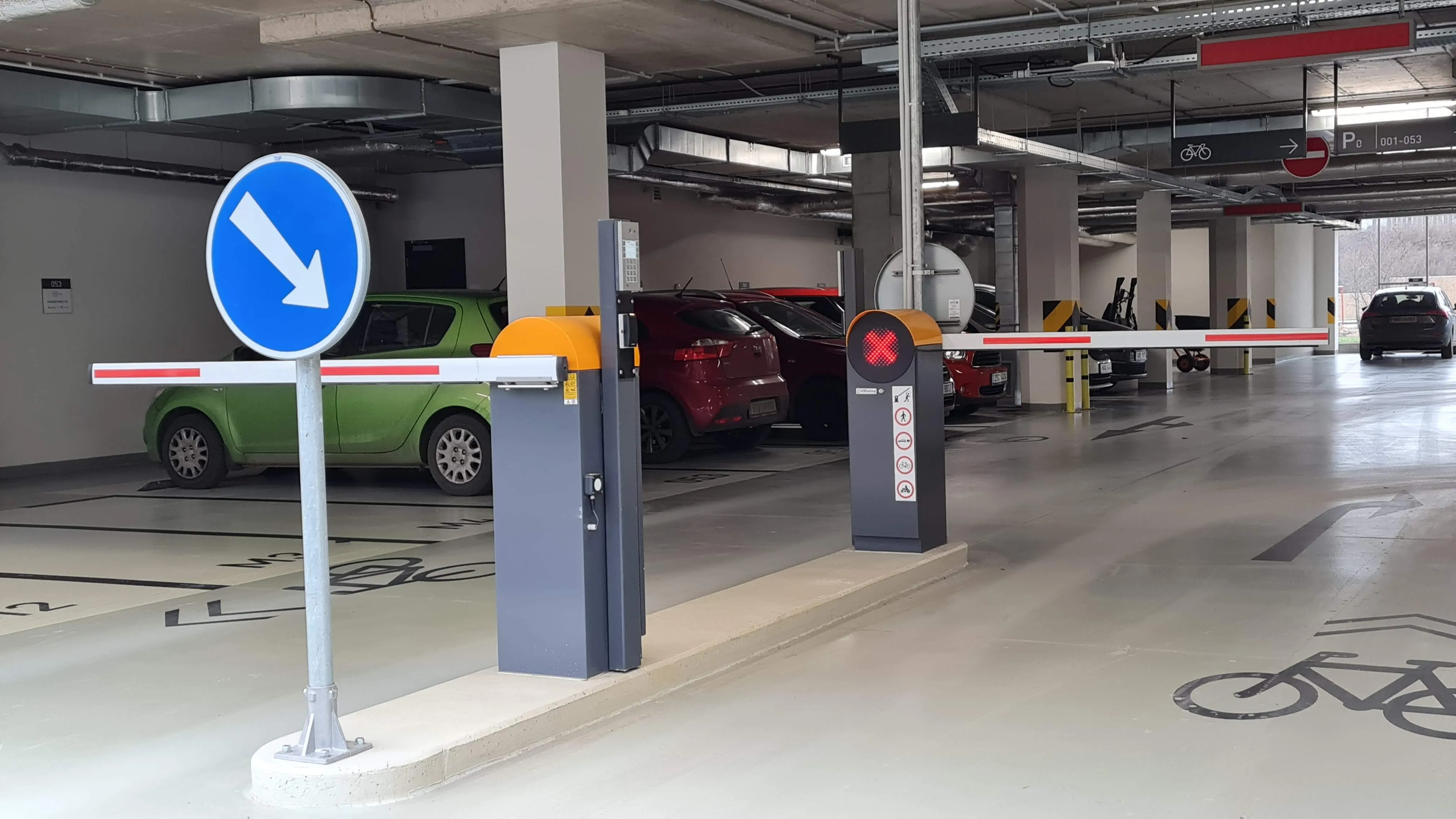 Vstupná časť podzemnej garáže s dvoma automatickými parkovacími závorami Park S v tmavosivej farbe s oranžovými detailmi a bielymi ramenami s červenými pruhmi, doplnené o modrú navigačnú šípku smerujúcu nadol a červené signalizačné svetlo, s vyznačenými cyklistickými pruhmi a zaparkovanými autami v pozadí.