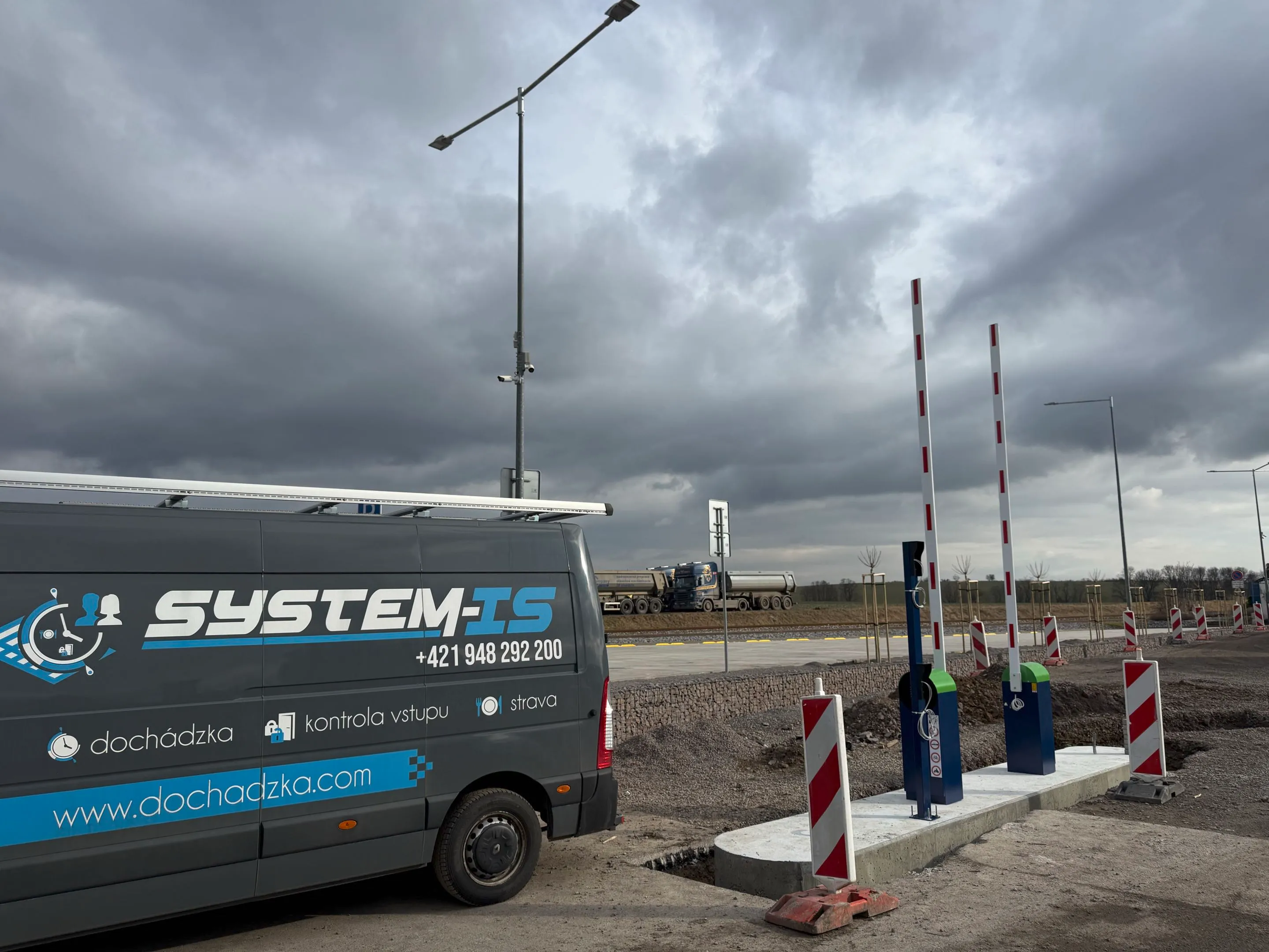 Inštalácia automatických parkovacích závor Park S s bielymi ramenami a červenými reflexnými pruhmi, vedľa dodávky "SYSTEM-IS" s logom dochádzka a kontrola vstupu, v exteriérovom prostredí pod zamračenou oblohou.