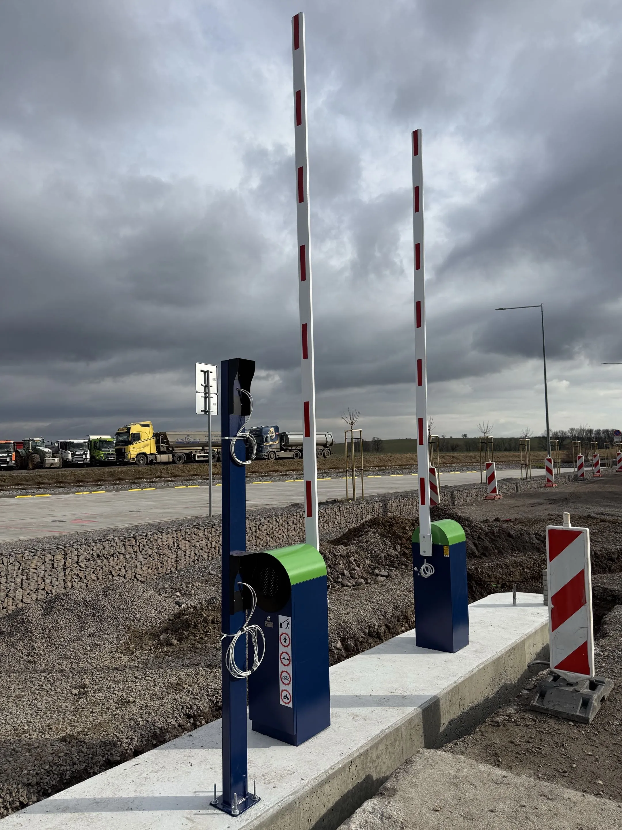 Dve automatické parkovacie závory Park S s tmavomodrými stĺpikmi a zelenými hornými krytmi, s bielymi ramenami s červenými reflexnými pruhmi v zdvihnutej polohe, inštalované na betónovom podklade pri štrkovej ceste s nákladnými vozidlami v pozadí, pod zamračenou oblohou.