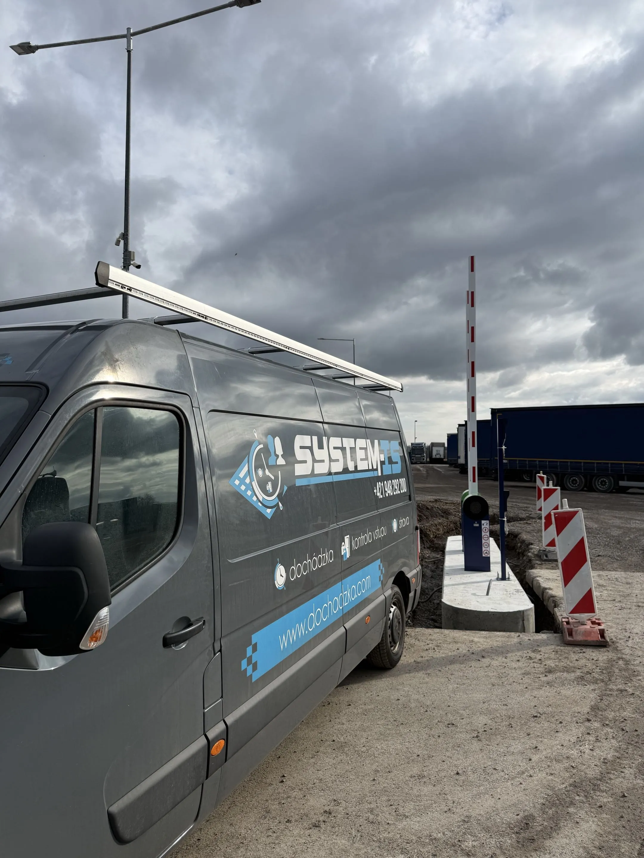 Automatická parkovacia závora Park S s tmavomodrým stĺpikom, zeleným horným krytom a bielym ramenom s červenými pruhmi, umiestnená pri dodávke "SYSTEM-IS", ktorá slúži na dochádzku a kontrolu vstupu, v exteriérovom prostredí pod zamračenou oblohou.